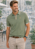 Hombre sonriente vistiendo un polo verde pálido de calidad premium con detalles en cuello y mangas, combinado con pantalones claros en ambiente urbano relajado.