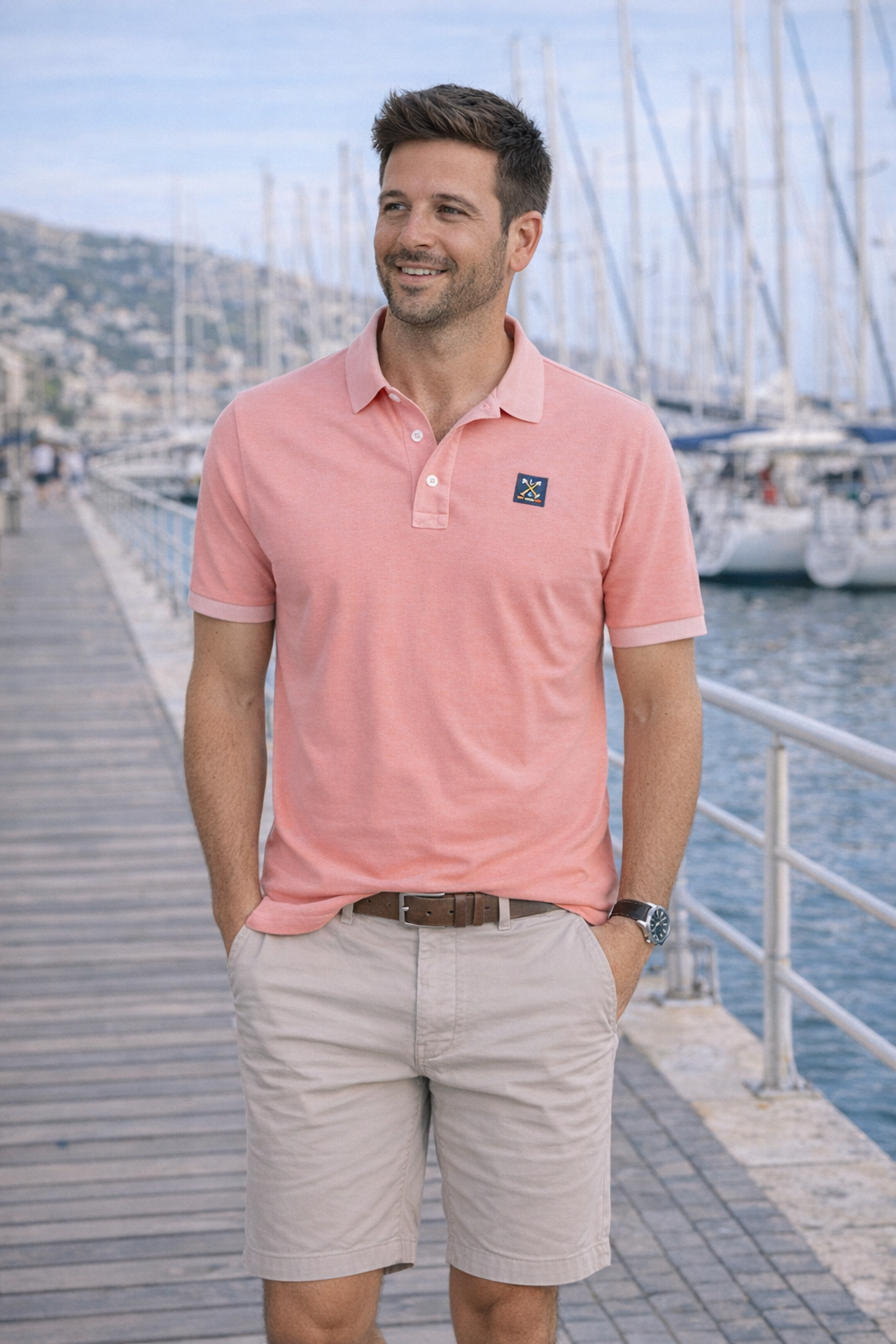 Hombre sonriente con polo color naranja coral de algodón y elastano, combinado con bermudas claras, posando en un muelle junto a un puerto deportivo.