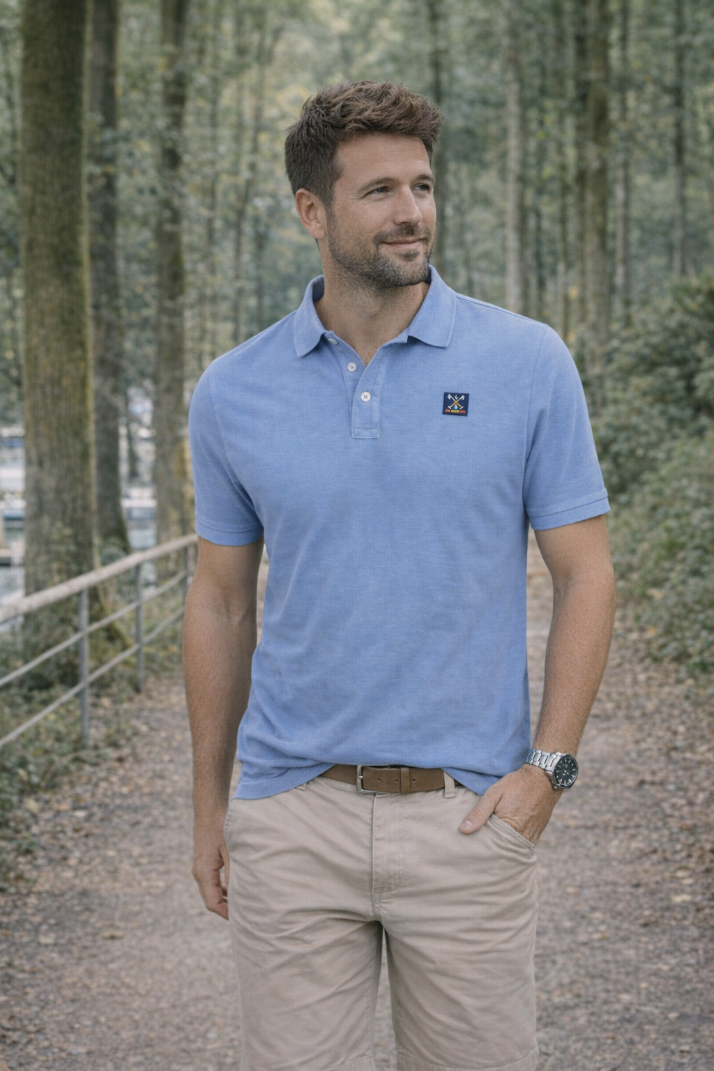 Hombre vestido con polo azul demil de algodón y elastano, modelo casual de moda masculina premium, posando en un entorno natural con árboles y sendero.