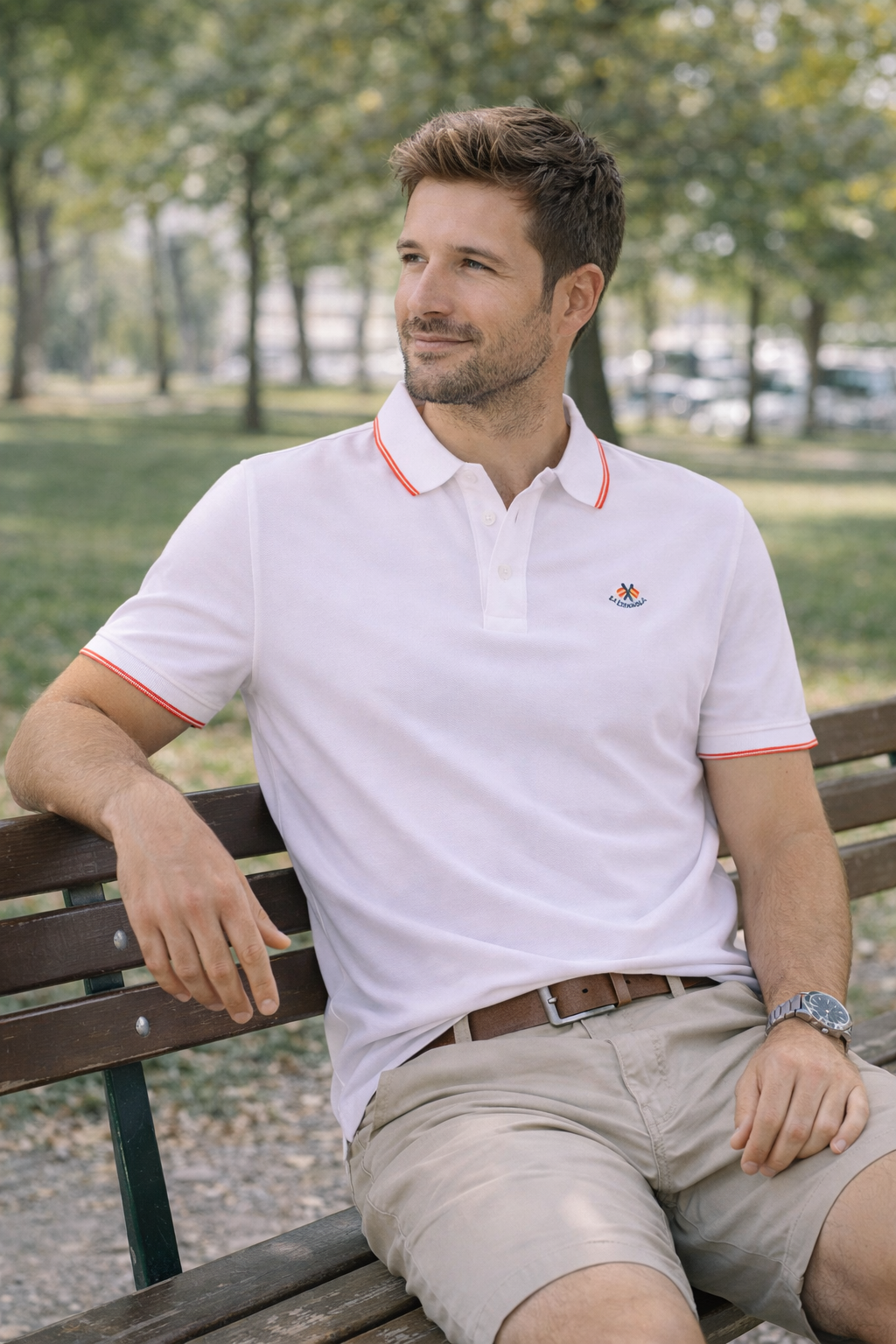Hombre sentado en banco de parque vistiendo polo blanco con detalles en rojo en el cuello y mangas, combinado con pantalones cortos beige, moda masculina premium con tejidos de algodón y elastano.